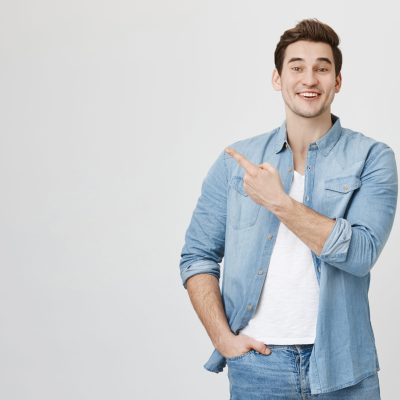 Studio portrait of attractive european man in denim glothes, pointing at upper left corner with index finger, expressing excitement, happiness and surprise, over gray background. Advertisement concept